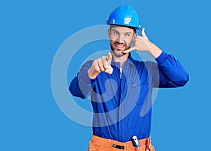 Young handsome man wearing worker uniform and hardhat smiling doing talking on the telephone gesture and pointing to you