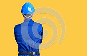 Young handsome man wearing worker uniform and hardhat standing backwards looking away with arms on body