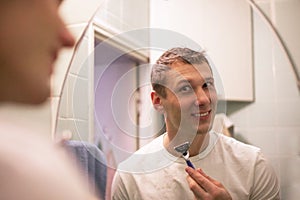 Young handsome man in white t-shirt shaves in front of bathroom mirror in the morning.