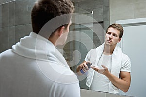 Young handsome man with white towel on his shoulders looking in the bathroom mirror putting shaving foam on his hand