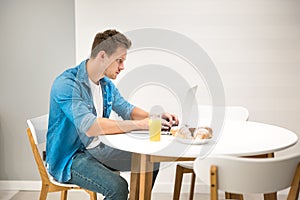 Young handsome man working in his laptop from home while having croissant and fresh orange juice for lunch looking