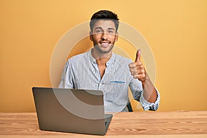 Young handsome man working at the office with laptop smiling happy and positive, thumb up doing excellent and approval sign