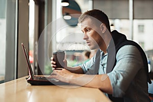 Young handsome man working remotely in his laptop holdscup of hot coffee in cafe looks surprised