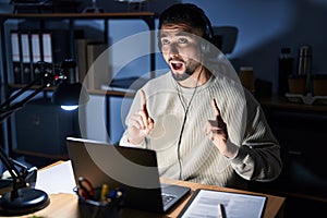 Young handsome man working using computer laptop at night amazed and surprised looking up and pointing with fingers and raised