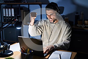 Young handsome man working using computer laptop at night angry and mad raising fist frustrated and furious while shouting with