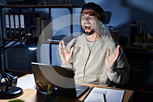 Young handsome man working using computer laptop at night crazy and mad shouting and yelling with aggressive expression and arms