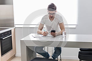 Young handsome man is writing something on paper notebook and holding mobile smart phone at home. business concept
