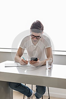 Young handsome man is writing something on paper notebook and holding mobile smart phone at home. business concept