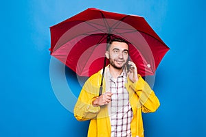 Young handsome bearded man in yellow raincoat with red umbrella talking by mobile phone isolated over blue background