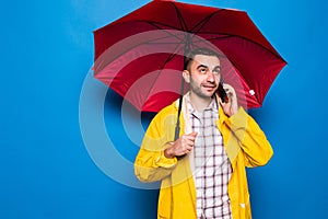 Young handsome bearded man in yellow raincoat with red umbrella talking by mobile phone isolated over blue background