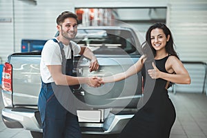 Young handsome mechanic and beautiful woman client shaking hands in gratitude for great service she shows like sign