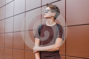 Young handsome modern hipster man in black stylish sunglasses in a stylish t-shirt in a black sports backpack near a wall