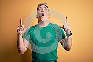 Young handsome modern man wearing casual green t-shirt over yellow background amazed and surprised looking up and pointing with