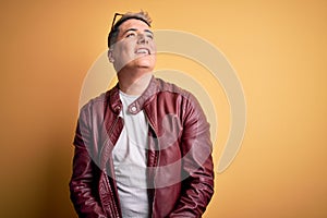 Young handsome modern man wearing fashion leather jacket and sunglasses over yellow background looking away to side with smile on