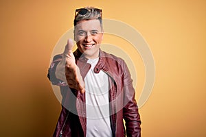 Young handsome modern man wearing fashion leather jacket and sunglasses over yellow background smiling friendly offering handshake
