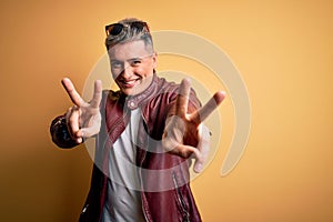 Young handsome modern man wearing fashion leather jacket and sunglasses over yellow background smiling looking to the camera