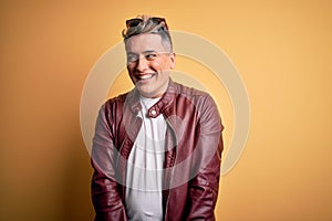 Young handsome modern man wearing fashion leather jacket and sunglasses over yellow background smiling looking to the side and