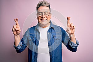 Young handsome modern man wearing glasses and denim jacket over pink isolated background gesturing finger crossed smiling with