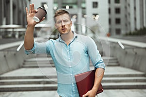 Young handsome office worker holding laptop and cup of coffee walking down the street near business center