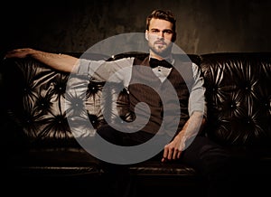 Young handsome old-fashioned bearded man sitting on comfortable leather sofa on dark background.