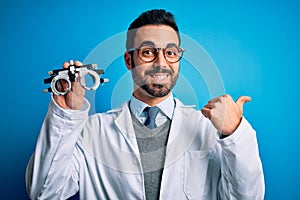 Young handsome optical man with beard holding optometry glasses over blue background Pointing to the back behind with hand and