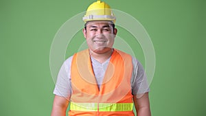 Young handsome overweight Asian man construction worker against green background
