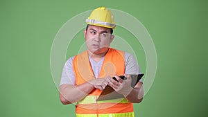 Young handsome overweight Asian man construction worker against green background