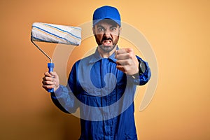 Young handsome painter man with beard wearing blue uniform and cap painting using roller annoyed and frustrated shouting with