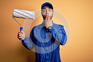 Young handsome painter man with beard wearing blue uniform and cap painting using roller cover mouth with hand shocked with shame