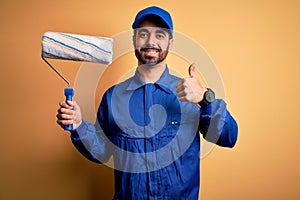 Young handsome painter man with beard wearing blue uniform and cap painting using roller happy with big smile doing ok sign, thumb