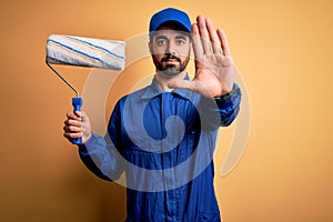 Young handsome painter man with beard wearing blue uniform and cap painting using roller with open hand doing stop sign with