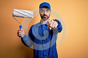 Young handsome painter man with beard wearing blue uniform and cap painting using roller pointing with finger to the camera and to