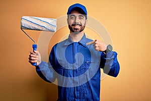 Young handsome painter man with beard wearing blue uniform and cap painting using roller with surprise face pointing finger to