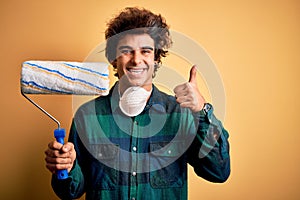 Young handsome painter man holding roller standing over isolated yellow background happy with big smile doing ok sign, thumb up