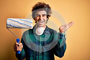 Young handsome painter man holding roller standing over isolated yellow background very happy and excited, winner expression