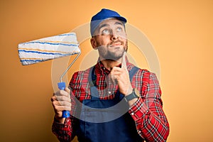 Young handsome painter man painting wall using roller over isolated yellow background serious face thinking about question, very