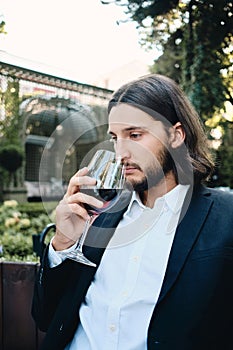 Young handsome pensive brunette bearded man thoughtfully drinking wine in restaurant outdoor