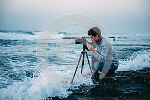 Young handsome photographer with curly hair, a gray sweater and jeans with a tripod and a camera on the beach, drenched in waves.