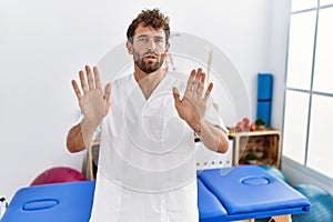 Young handsome physiotherapist man working at pain recovery clinic moving away hands palms showing refusal and denial with afraid