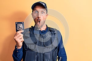 Young handsome policeman wearing uniform and bulletprof holding police badge scared and amazed with open mouth for surprise,