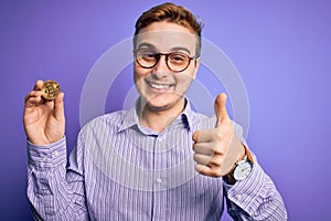 Young handsome redhead businessman holding cryptocurrency bitcoin over purple background Smiling happy and positive, thumb up