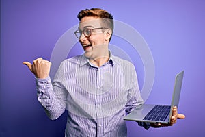 Young handsome redhead businessman working using laptop over purple background pointing and showing with thumb up to the side with