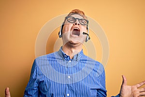 Young handsome redhead call center agent man wearing glasses working using headset crazy and mad shouting and yelling with