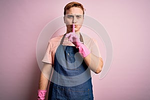 Young handsome redhead cleaner man doing housework wearing apron and gloves asking to be quiet with finger on lips