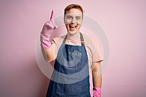 Young handsome redhead cleaner man doing housework wearing apron and gloves pointing finger up with successful idea