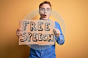 Young handsome redhead man asking for freedom holding banner with free speech message scared and amazed with open mouth for