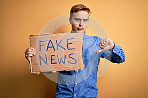 Young handsome redhead man holding banner with fake news message over yellow background with angry face, negative sign showing