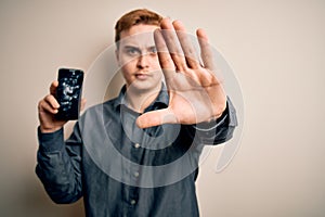 Young handsome redhead man holding broken smartphone showing cracked screen with open hand doing stop sign with serious and