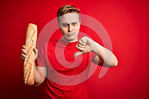 Young handsome redhead man holding fresh homemade bread over isolated red background with angry face, negative sign showing