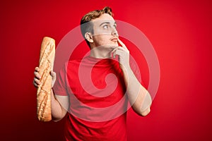 Young handsome redhead man holding fresh homemade bread over isolated red background serious face thinking about question with